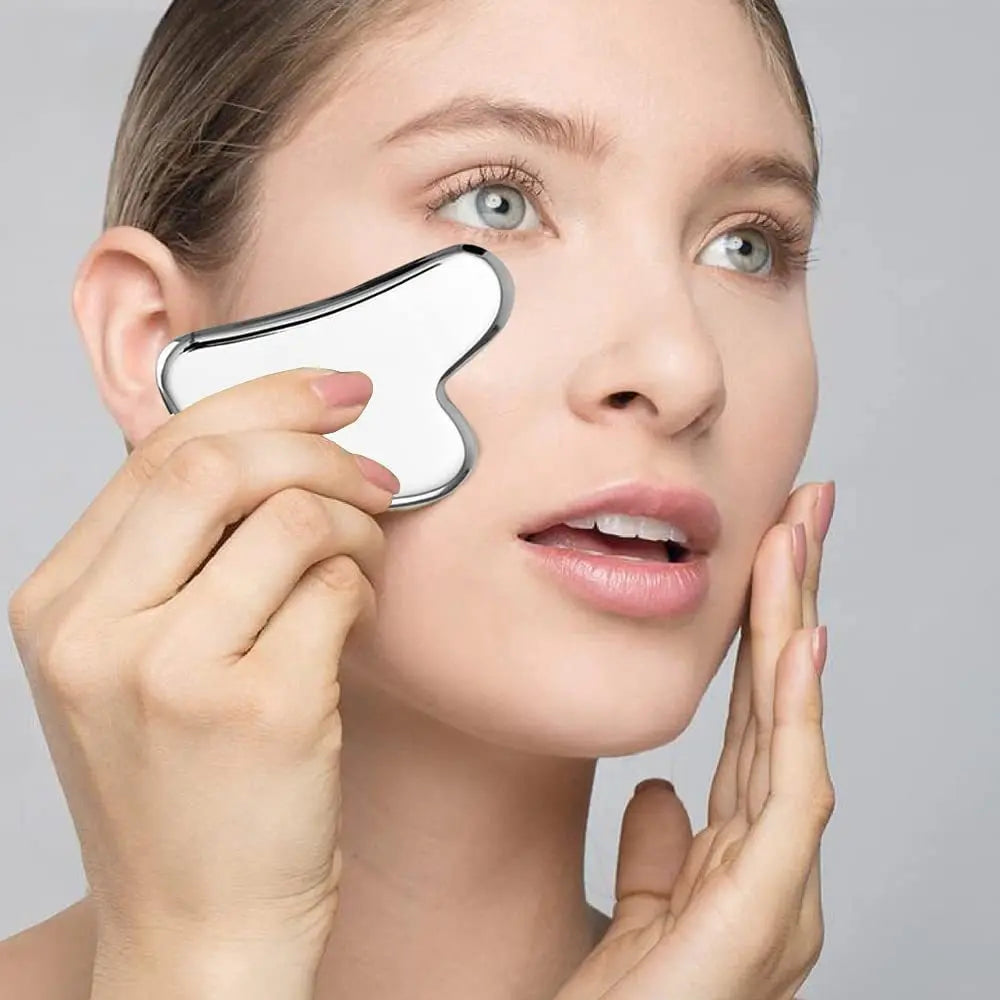 A woman is using a stainless steel gua sha tool on her face for facial massage.