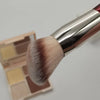 Un pinceau de maquillage avec une ferule argentée et un poil synthétique dense rose pâle et brun, à côté de son emballage.