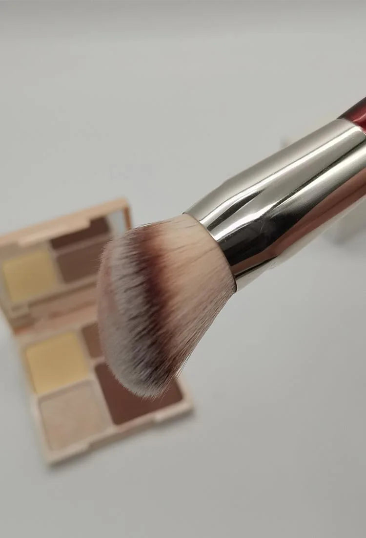 Un pinceau de maquillage avec une ferule argentée et un poil synthétique dense rose pâle et brun, à côté de son emballage.