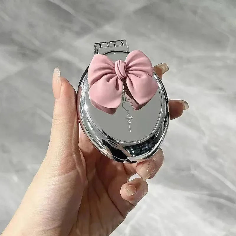 A hand holds a silver folding massage comb with a pink bow on a circular mirror.