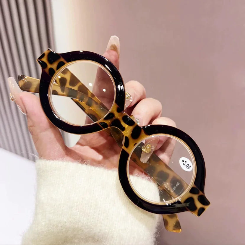 A hand holds a pair of round tortoiseshell acetate reading glasses with anti-blue light lenses.