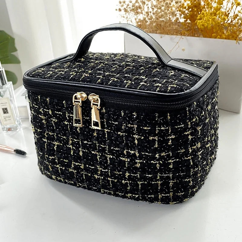 A black knitted canvas makeup pouch with a gold zipper and handle resting on a white surface.