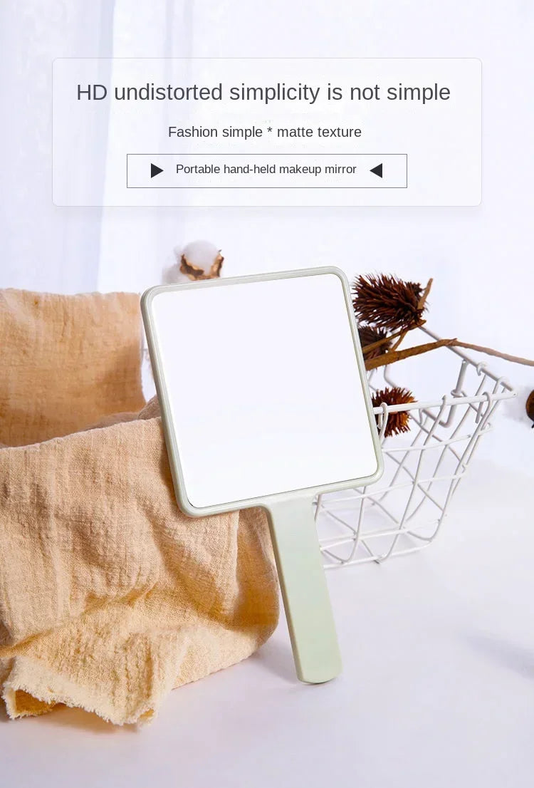 Un miroir de maquillage rectangulaire vert clair à main, à côté d'un panier tissé et d'une serviette beige.