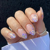 A close-up of a hand displaying press-on acrylic flower nails with purple and white floral accents.