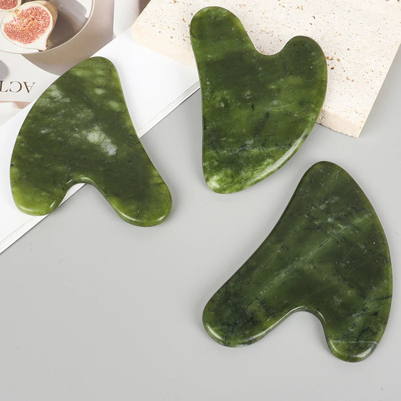 Three smooth green jade gua sha tools are displayed on a white surface next to a piece of natural stone.