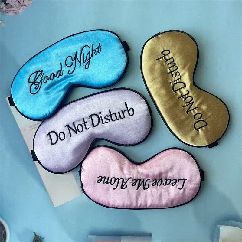 Three embroidered silk sleep masks in blue, gold, and pink are displayed on a light blue background.