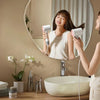 A woman is using a professional high-speed hair dryer in front of a mirror while sitting by a bathtub.