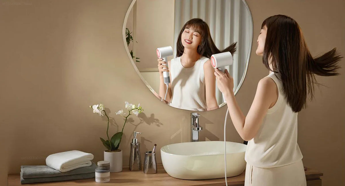 A woman is using a professional high-speed hair dryer in front of a mirror while sitting by a bathtub.