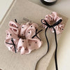 Scrunchies en satin à pois roses, attachés avec un ruban noir, sur un tissu beige.