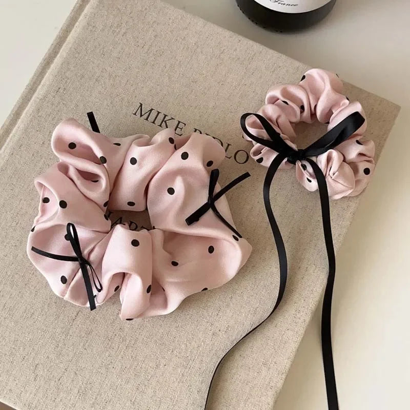 Scrunchies en satin à pois roses, attachés avec un ruban noir, sur un tissu beige.