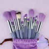 A collection of thirteen professional makeup brushes in various shades of purple and gray displayed on a lavender cloth.