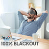 A man wearing a black sleep mask is resting his head on his hands while working at a desk.
