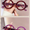 A pair of purple-tinted round acetate reading glasses with a leopard print design being held by a hand.