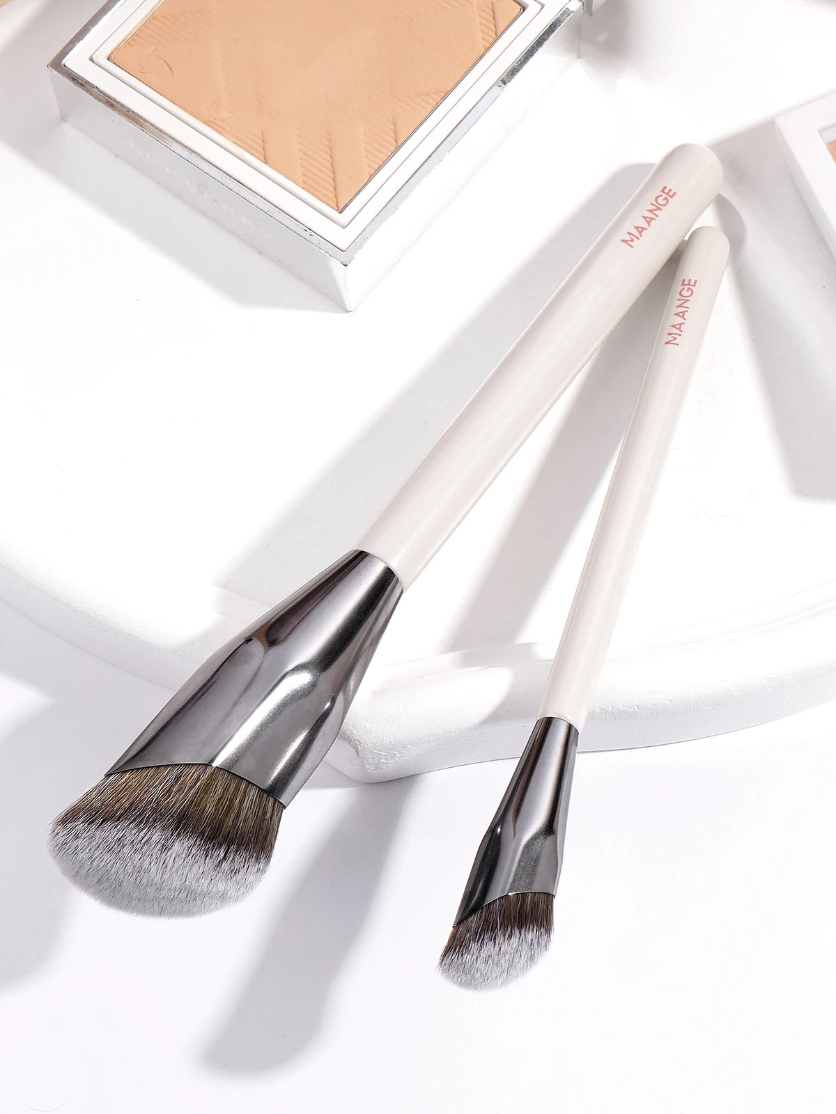 A pair of beige makeup brushes with silver ferrules and fluffy bristles are displayed on a white surface.
