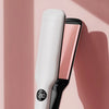 A white and pink hair straightener is shown vertically against a soft pink background.