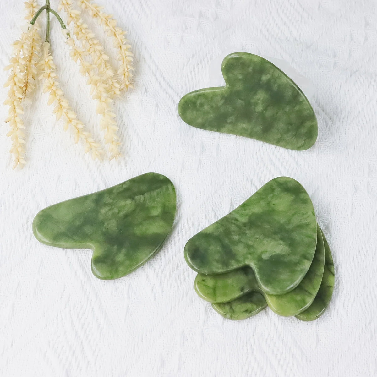 Green jade gua sha tools shaped like hearts resting on a white surface next to a sprig of dried flowers.