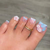 A hand displaying press-on fake nails with light pink and blue floral designs and a gold band ring.