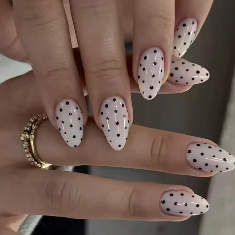 Close-up of a hand displaying reusable press-on nails with a white base and black polka dots on an almond shape.