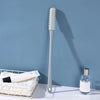 A silver long-handled manual foot scrubber brush is displayed against a blue wall next to a white product box.