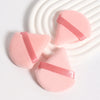 A set of pink triangular cosmetic powder puffs with pink ribbon accents resting on a white striped surface.