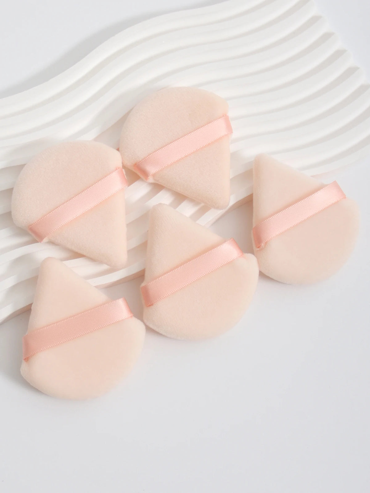 A set of three pink cosmetic powder puffs with light pink ribbon accents resting on a white striped surface.