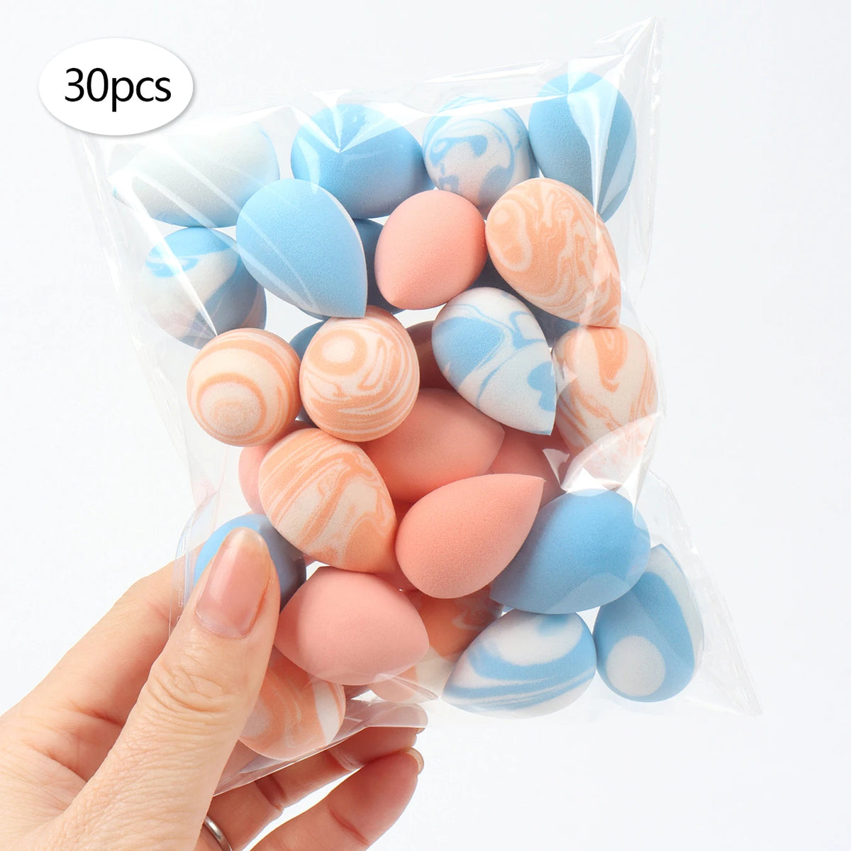 A hand holds a clear bag filled with 30 reusable expandable makeup sponges in various pastel blue and peach colors.