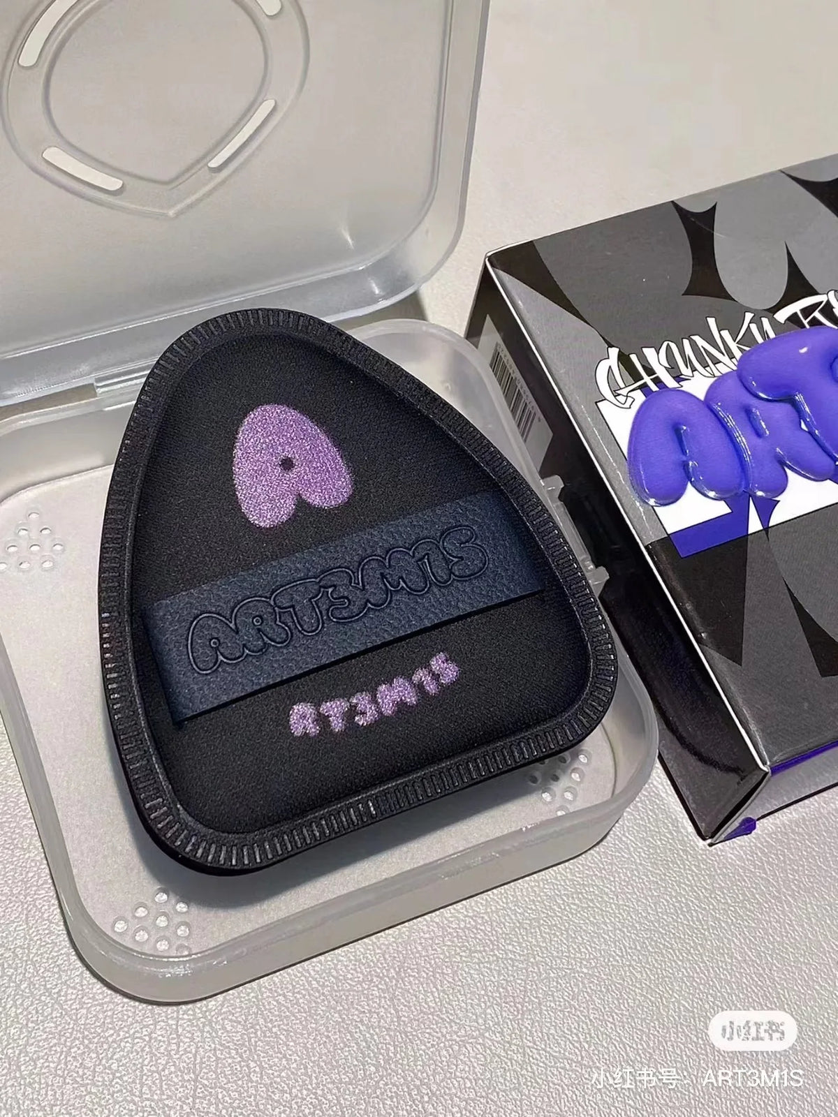 Un applicateur cosmétique noir à flockage avec des lettres violettes, présenté dans un contenant en plastique transparent.