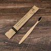 A bamboo toothbrush packaged in natural paper, lying next to the wooden brush on a dark brown surface.