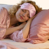 Une femme aux cheveux blonds dort paisiblement, portant un bandeau de soie rose, sur un oreiller rose.