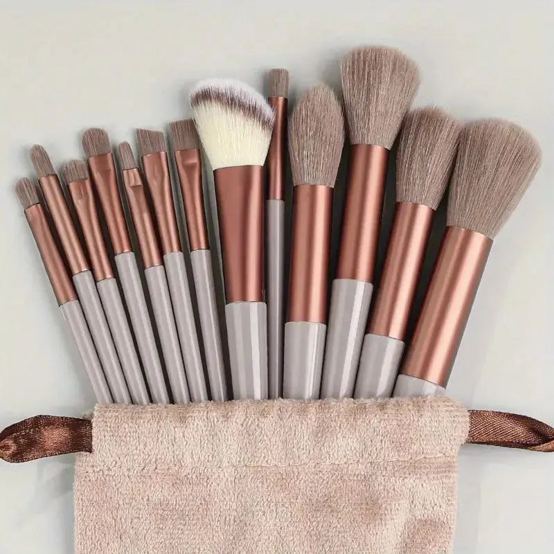 A set of thirteen professional makeup brushes with rose gold and gray handles resting on a beige towel.