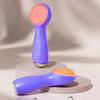 A purple vibrating LED facial cleansing brush is shown on a light gray surface.