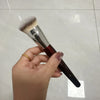 Une main tient un pinceau de maquillage liquide angulé avec un anneau doré sur fond blanc.