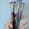 Une main tient trois pinceaux de maquillage liquide inclinés avec des ferrules en argent et des poils synthétiques gris.