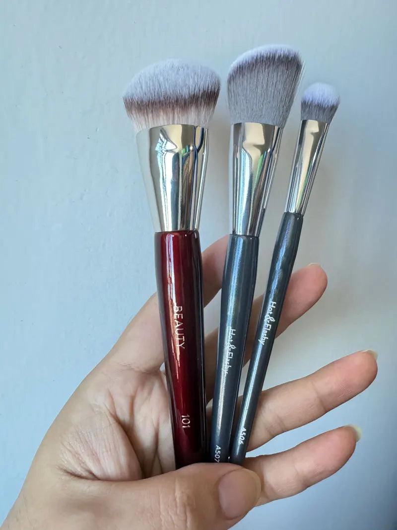 Une main tient trois pinceaux de maquillage pour fond de teint anglés avec des ferrules en argent et des poils synthétiques g