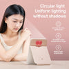 A close-up of a foldable LED makeup mirror with a light pink frame and a red accent, shown on a table.