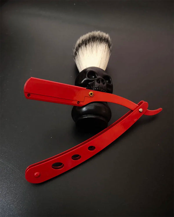 A red straight edge barbershop razor set with a black skull and silver bristles is displayed on a dark surface.