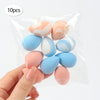 A hand holds a clear plastic bag containing ten reusable expandable makeup sponges in various pastel colors like pink, peach,