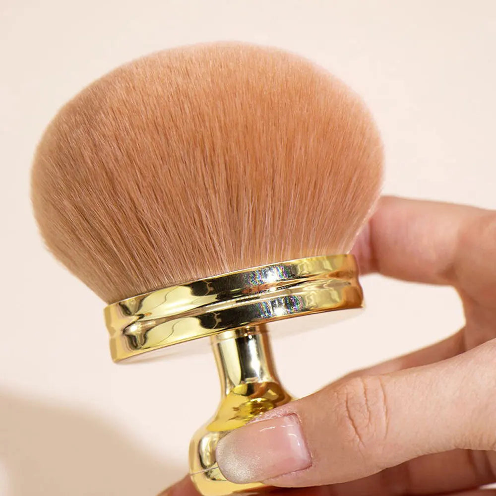 Un grand pinceau de maquillage poudre, couleur pêche clair et manche en métal doré, tenu dans une main.