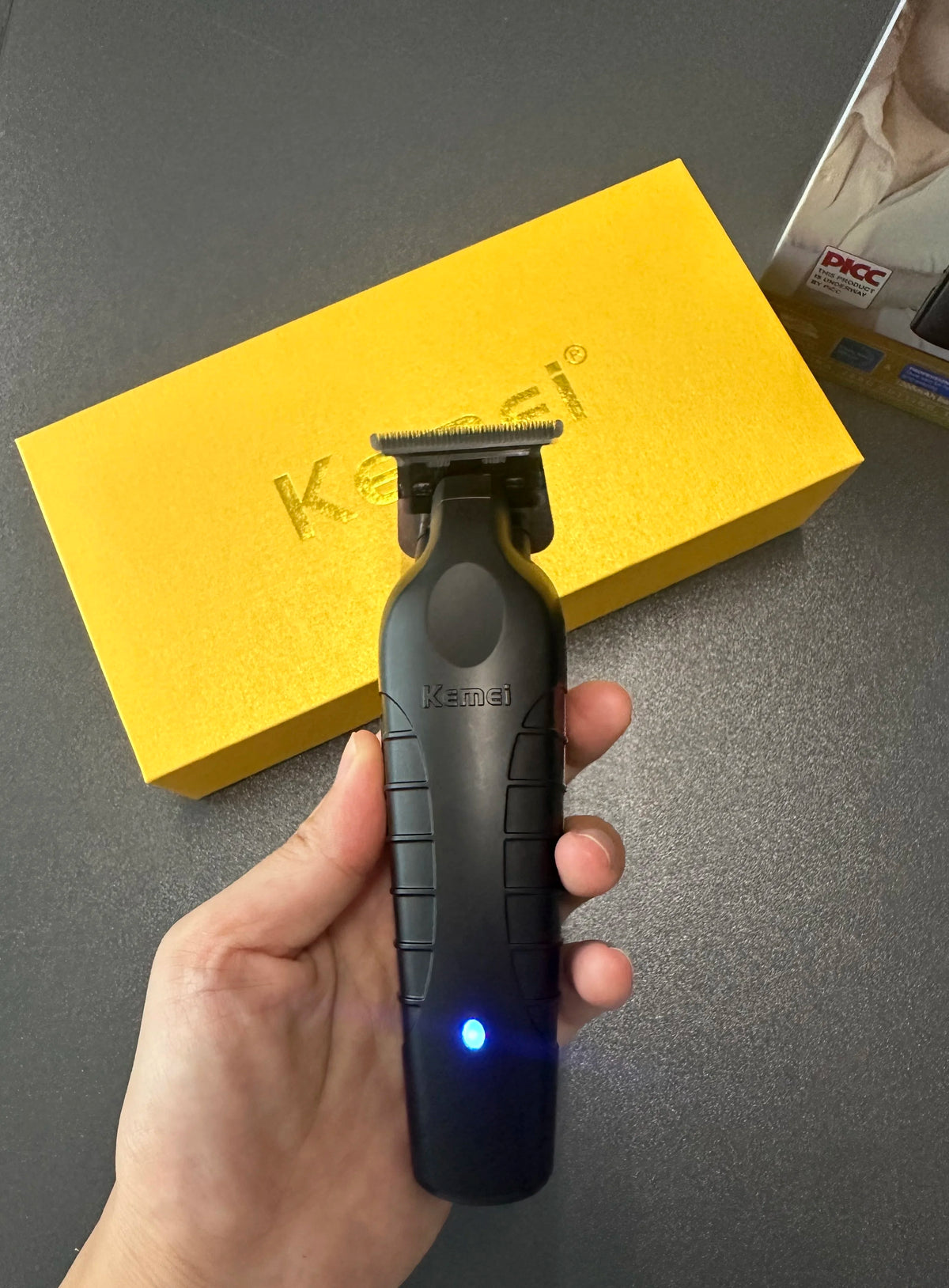 A hand holds a black cordless electric hair clipper against a yellow box.
