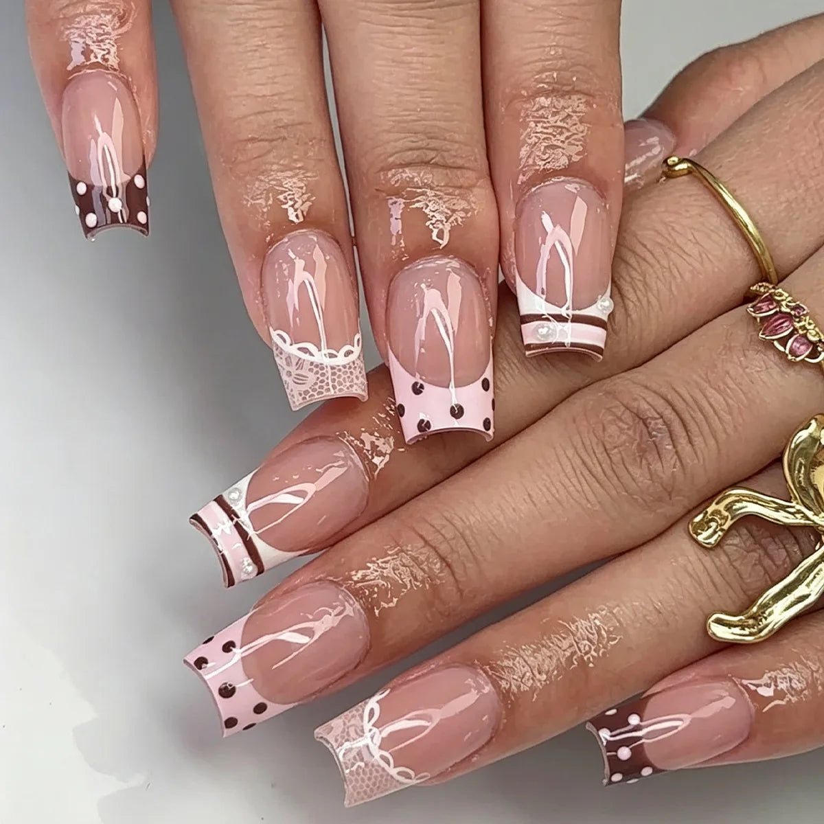 A close-up of a hand displaying gothic Y2K press-on nails with white lace and black dot designs on a sheer pink background.