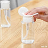 A hand is dispensing liquid from a clear refillable push-down pump dispenser bottle onto a wooden surface.