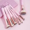 A set of seven pink and gold makeup brushes displayed on a pink background.