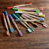 A collection of ten eco-friendly bamboo toothbrushes in various colors and bristles laid out on a wooden surface.