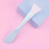 A light blue silicone facial mask brush with a pink head is shown resting on a purple box.