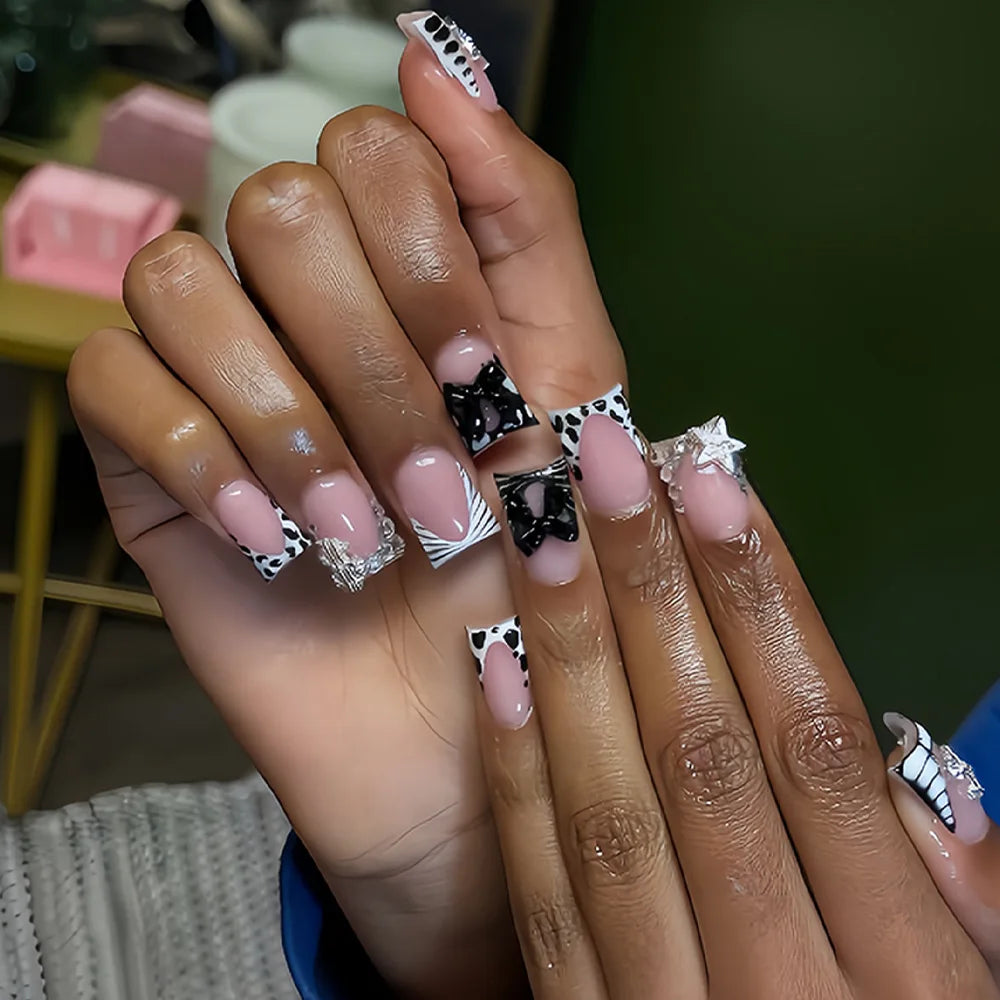 A close-up of a hand displaying nude pink press-on nails decorated with leopard print and silver 3D charms.