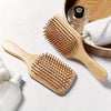 A wooden bamboo scalp and hair growth comb resting on a white surface next to a wooden bowl and a white towel.