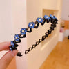 A black rhinestone wave headband with blue flowers is held by a hand against a light wall.
