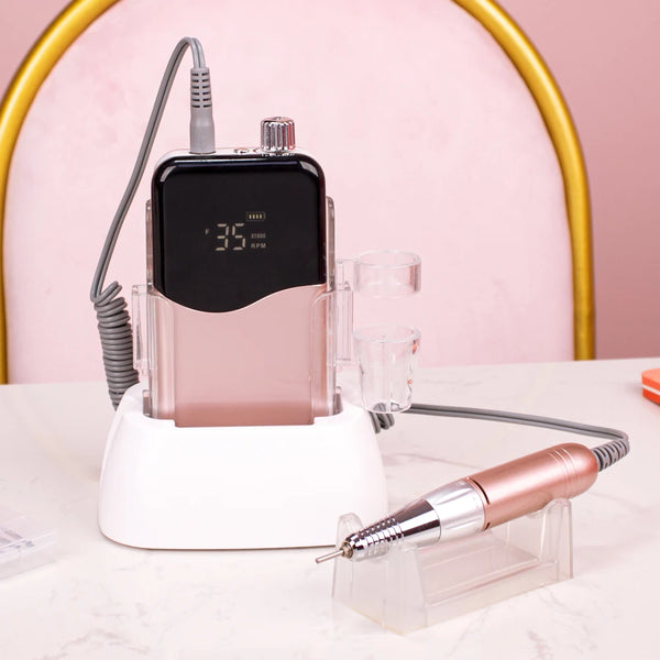 A cordless rechargeable nail drill device with a pink body and black battery, resting on a white base.