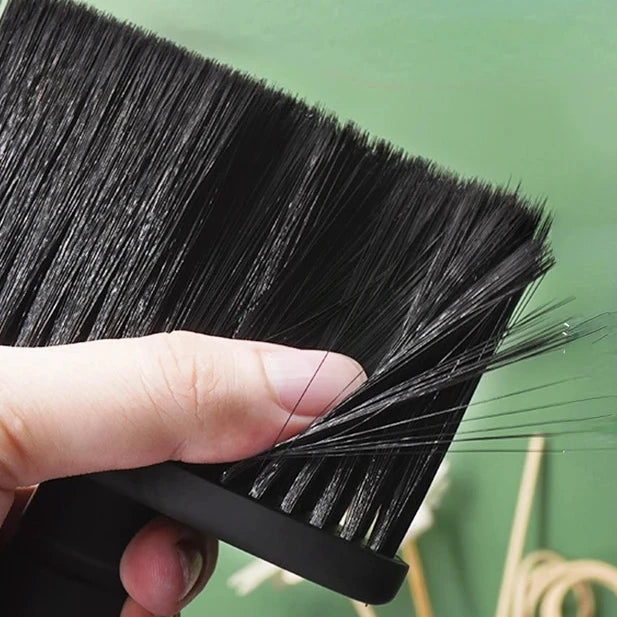 A hand is holding a black soft bristle hair duster brush, showing the dense bristles and handle.