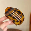 A hand holds an oval flat hair clip with a leopard print pattern in shades of amber and black.